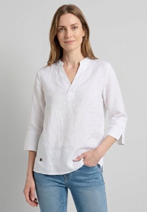 Mujer con cabello castaño claro que lleva una blusa de lino blanca con cuello en V y mangas tres cuartos, y vaqueros azules, de pie frente a un fondo gris.