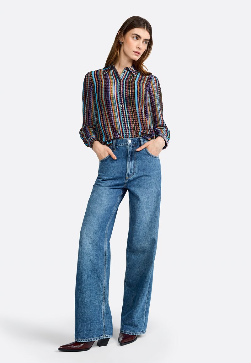 Blusa a righe multicolore con colletto, abbinata a jeans a palazzo blu in denim a vita alta. La modella indossa dei tacchi a punta.