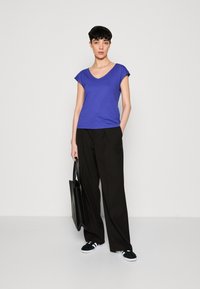Lila V-necka T-shirt s krátkými rukávy, spárovaný s černými kalhotami s širokými nohavicemi, černou taškou přes rameno a černými teniskami. Hladký povrch.