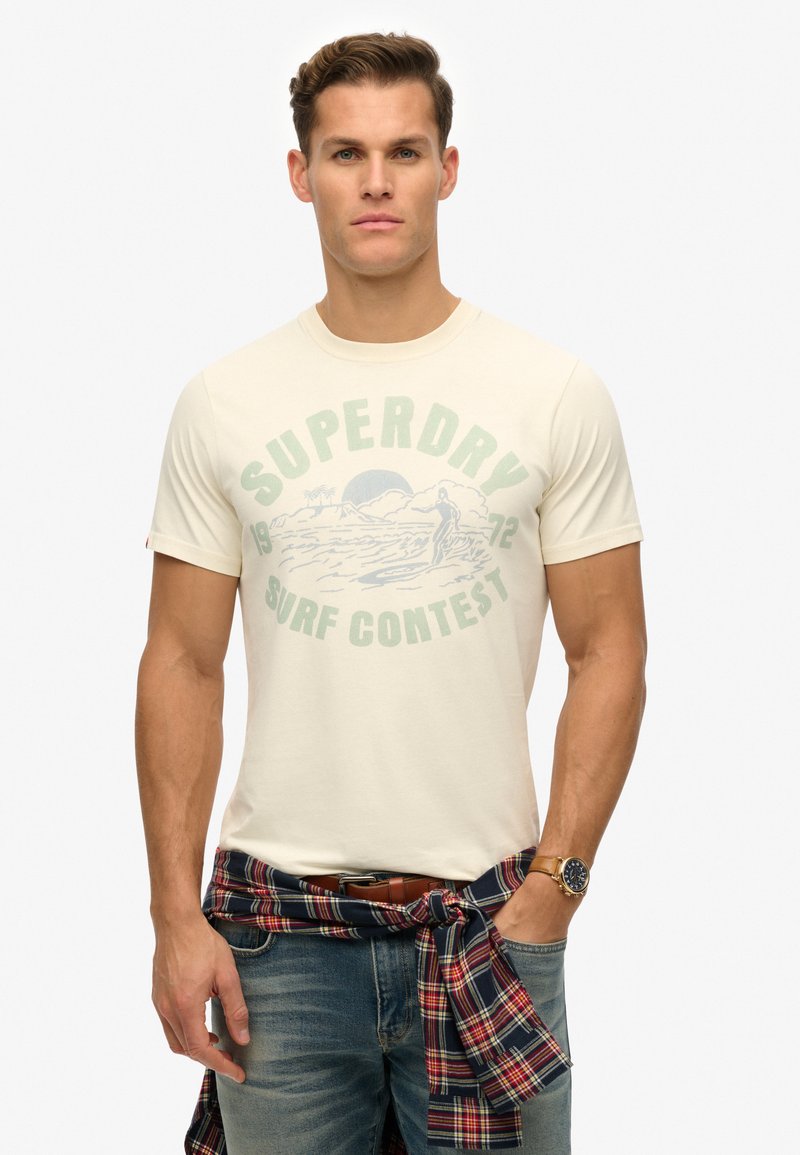 Camiseta de color crema con un gráfico de "SUPERDRY SURF CONTEST" en verde y azul. Llevada con una camisa de cuadros atada a la cintura.