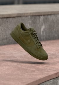 Πράσινο ελιάς αθλητικό παπούτσι Nike που αιωρείται στον αέρα πάνω από σκυρόδεμα, με φόντο έναν τοίχο από υφή γκρι πέτρας.