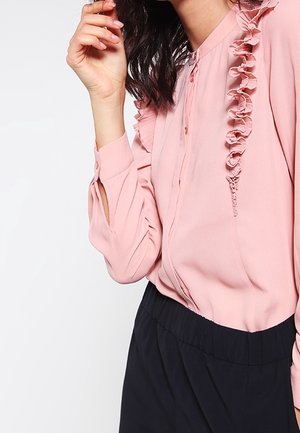 Nainen, joka pukeutunut vaaleanpunaiseen pitkähihnaiseen puseroon, jossa on röyhelöyksityiskohtia, ja mustiin korkeavyötäröisiin housuihin, käsi kohotettuna lähelle kasvoja.