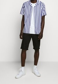 Camicia a manica corta con bottoni, a righe verticali viola e bianche, abbinata a pantaloncini neri arrotolati e sneakers alte bianche.