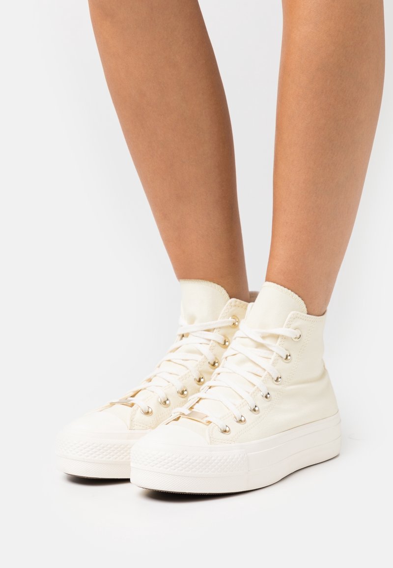 Jambes portant des baskets montantes blanches en toile avec des semelles épaisses et des lacets blancs sur un fond clair uni.