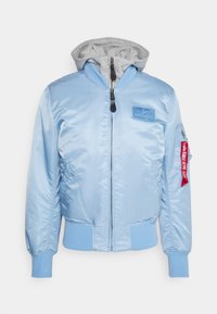 Lichtblauwe bomberjas met geribde manchetten en zoom, grijze afneembare capuchon, ritssluiting aan de voorkant, borstpatch en rode “Remove Before Flight” label op het mouwzakje.