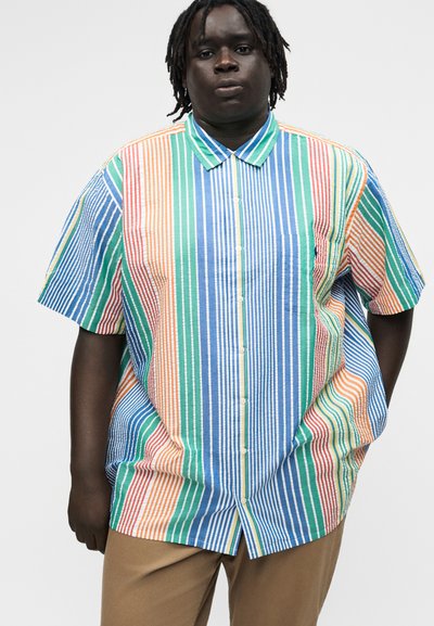 Άτομο που φοράει πουκάμισο με κοντά μανίκια, κουμπωτό, με κάθετες πολύχρωμες ρίγες και μπεζ παντελόνι σε απλό φόντο.