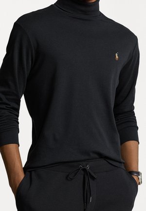 Mężczyzna ubrany w czarną, długorękawową bluzkę z golfem z małym haftowanym logo zawodnika polo oraz czarne spodnie ze sznurkiem, z rękami w kieszeniach.