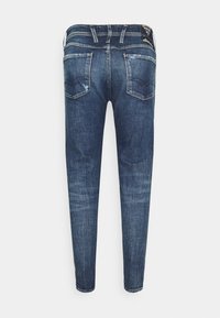 Bakvy av slim-fit blå denimjeans med slitna detaljer, bakfickor, bältesömmar och en svart etikett på midjan.
