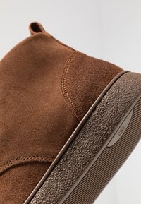 Chaussure en daim marron avec une semelle en caoutchouc marron texturée et des coutures visibles, présentée en angle pour montrer les détails latéraux et inférieurs sur un fond uni.