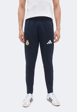 Homme portant un pantalon de sport bleu marine avec les logos du Real Madrid et d'Adidas, assorti à des baskets blanches Adidas.