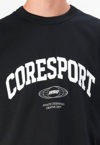 Svart bomulls t-skjorte med stor hvit tekst "CORESPORT" over et globusgrafikk og mindre tekst "ATHLETIC ESSENTIALS CREATIVE DEPT."