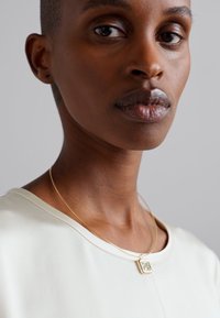 Collana d'oro con pendente quadrato, indossata sopra una maglietta liscia di colore chiaro. La pelle è di un marrone scuro e lo sfondo è neutro.