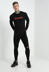 Camisa negra de manga larga con el texto rojo "THUGGER" en el frente. Jeans skinny negros rasgados y zapatillas negras. Tatuaje visible en el cuello.