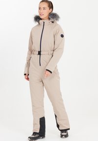 Beige vinteroverall med pälsfodrad huva, dragkedja fram, bälte i midjan, svarta detaljer vid ärmar och nedre ben, samt vattenavvisande tyg.