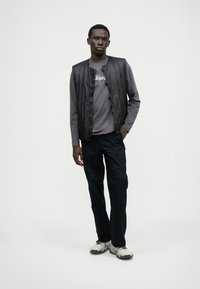 Μαύρο ελαφρύ γιλέκο πάνω από γκρι μακρυμάνικο πουκάμισο, συνδυασμένο με μαύρο παντελόνι και γκρι αθλητικά παπούτσια. Απλός σχεδιασμός με χαλαρή εφαρμογή.