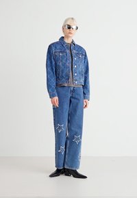 Jeans jakna s kvadratnim vzorcem in sprednjimi žepek, v kombinaciji s širokimi jeans hlačami z aplikacijami zvezd. Oboje v modrem denimu. Črni škornji in sončna očala.