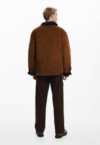 Braune Cordjacke mit blauer Kragen, lässiger Schnitt und strukturiertem Material. Getragen über dunkelbraunen Cargo-Hosen und robusten Schuhen.