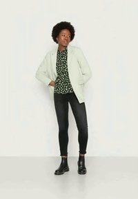 Blazer cinza claro sobre uma blusa estampada em preto e verde, combinado com jeans escuros e botas de tornozelo pretas, posicionado contra um fundo neutro.
