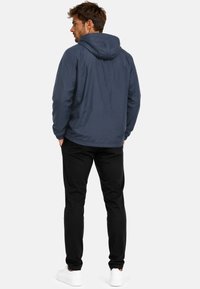 Homme debout de dos, portant une veste à capuche bleu marine, un pantalon noir et des baskets blanches sur un fond clair uni.