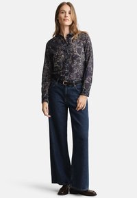 Schwarzes Hemd mit floralem Muster und Knöpfen, langen Ärmeln, kombiniert mit weit geschnittenen, dunkelblauen Jeans und braunen Lederschuhen.