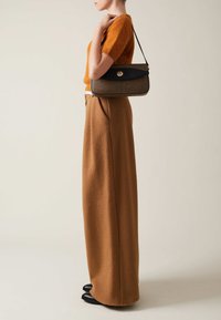 Borsa a tracolla marrone con texture, con una patta blu navy e una chiusura circolare. Il modello indossa un corto maglione arancione peloso e pantaloni larghi marroni.