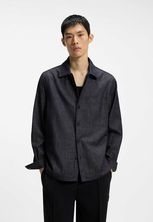 Jeune homme aux cheveux noirs courts porte une chemise gris foncé à boutons par-dessus un haut noir, debout les mains dans les poches sur fond blanc.