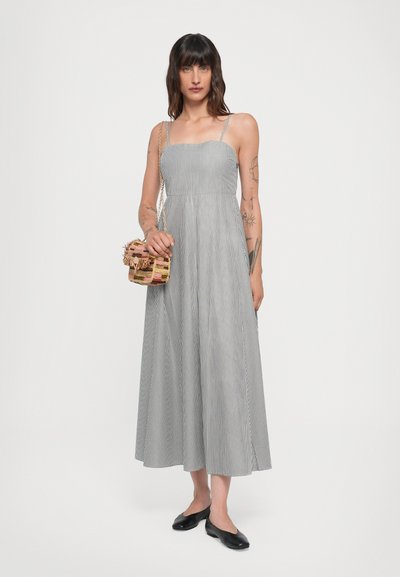 Gestreiftes, ärmelloses Midikleid in Schwarz und Weiß mit körperbetontem Oberteil und schwingendem Rock. Das Modell hält eine strukturierte, mehrfarbige Tasche.