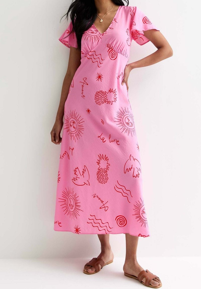 Robe maxi rose avec un col en V et des manches courtes volantées, ornée de motifs rouges représentant des ananas, des soleils et des dessins abstraits. Sandales plates.