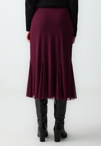 Jupe midi bordeaux avec des plis �à l'ourlet et des accents transparents, associée à des bottes en cuir noires montantes. Texture de tissu lisse.