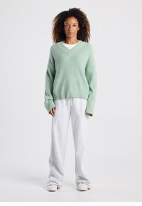 Pull en maille côtelée vert clair avec un col en V, associé à un pantalon de survêtement gris ample. La tenue présente une coupe décontractée et un look casual.