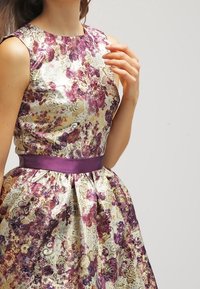 Nainen, jolla on yllään hihaton, kukallinen brokadimekko violetein, kulta- ja valkoisin kuvioin sekä violetti satiinivyö, käsi kohotettuna lähellä olkapäätä.