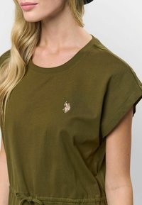 T-shirt scurt, de culoare verde măslinie, din bumbac, cu guler rotund și un mic logo brodat aurit pe piept.