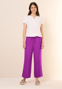 Witte korte mouw top met knoopjes henley detail, gecombineerd met losse, hoge taille, fel paarse broek en metalen sandalen.