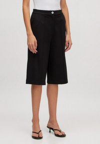 Culottes noirs avec une taille mi-haute, plis sur le devant et fermeture à un bouton. Les matériaux semblent lisses ; présente une silhouette décontractée et large.
