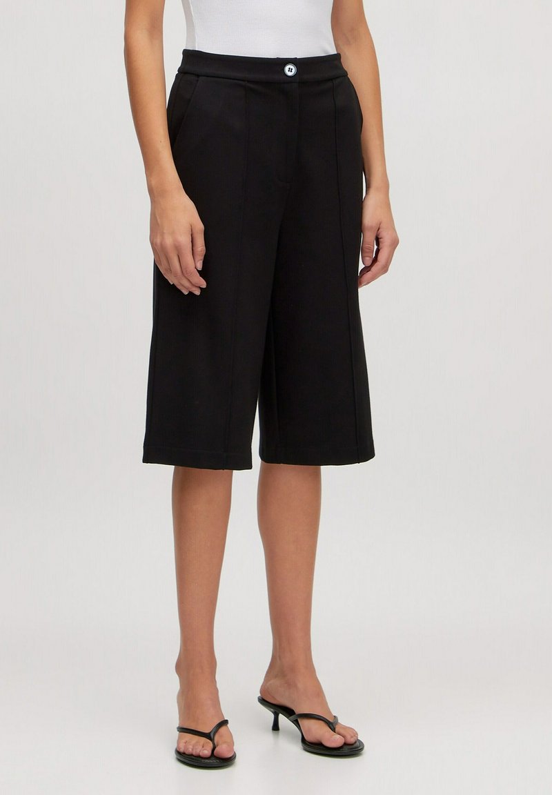 Culottes noirs avec une taille mi-haute, plis sur le devant et fermeture à un bouton. Les matériaux semblent lisses ; présente une silhouette décontractée et large.