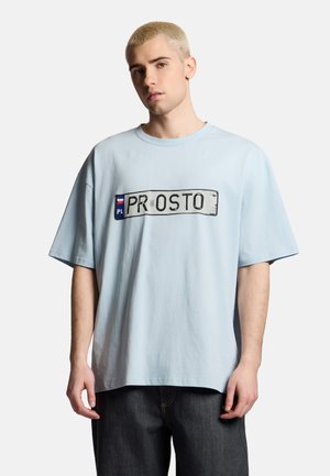 Młody mężczyzna o krótkich blond włosach, ubrany w jasnoniebieski oversize T-shirt z grafiką tablicy rejestracyjnej "PL PROSTO" i ciemne spodnie.