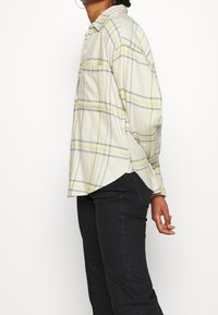 Person wearing a cream plaid button-up shirt with gray and yellow stripes, paired with black fitted pants, standing against a white background.