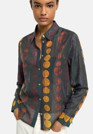 Kobieta ubrana w ciemnozieloną koszulę na guziki z dużymi żółtymi, fioletowymi i brązowymi okrągłymi wzorami oraz białe spodnie, z rękami w kieszeniach.