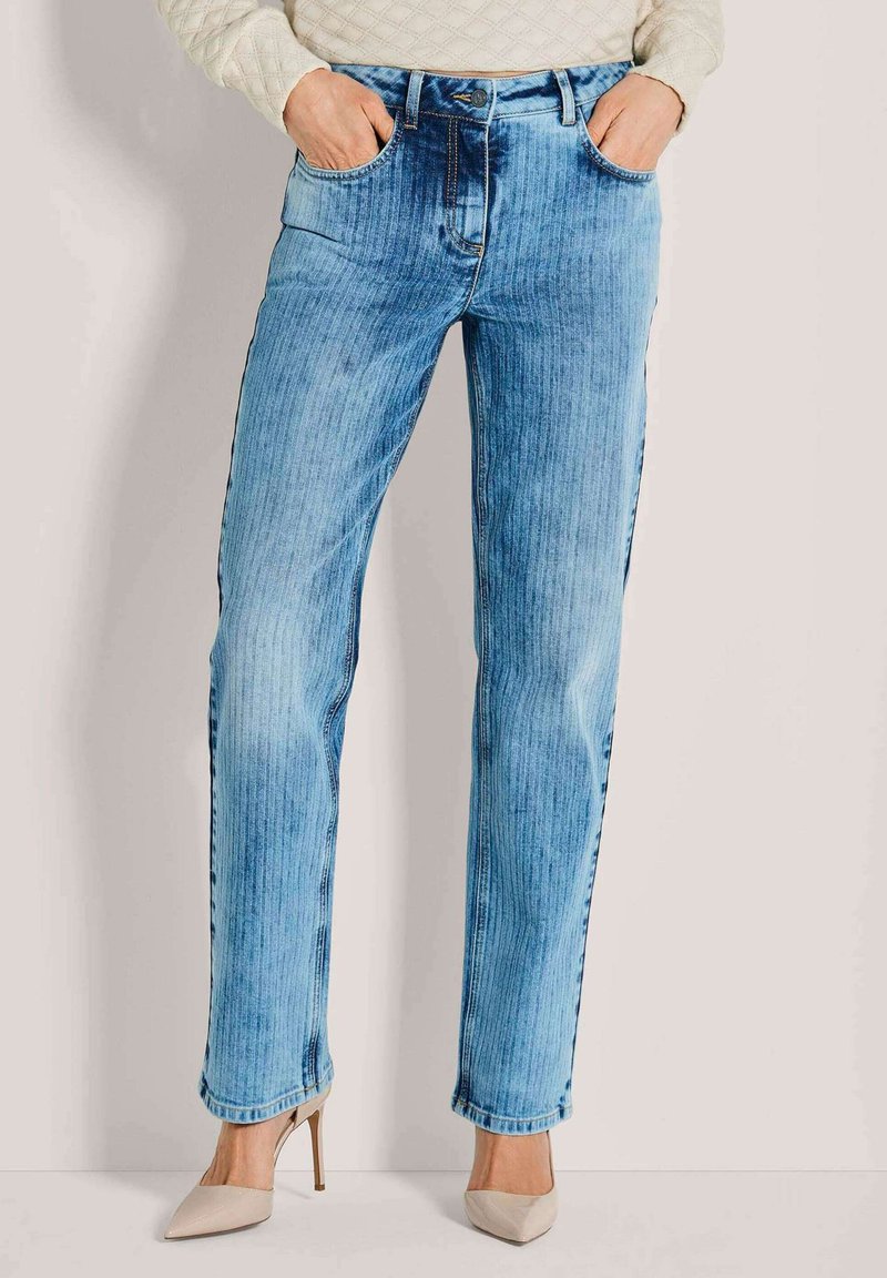 Frau trägt hellblaue, gerade geschnittene Jeans mit vertikalen Streifen und beigefarbene High-Heels, Hände in den Taschen, vor einem einfarbigen Hintergrund.