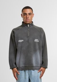 Pullover in pile grigio con colletto alto, zip a metà e maniche raglan. Presenta loghi stampati sul davanti e una texture leggermente sbiadita.