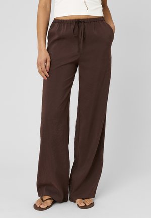 Femme portant un pantalon ample marron foncé à cordon, un top court blanc et des tongs marron, debout avec les mains dans les poches.