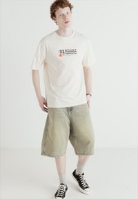 Crèmefarvet T-shirt med en sort og rød grafisk tryk, parret med lys grøn denimshorts og sorte sneakers med hvide såler.