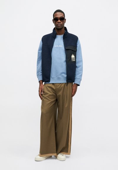 Námornícky fleece vest over svetlomodrou mikinou, hnedé široké nohavice s bočnými pruhmi a biele tenisky. Hladké textúry a vrstvený dizajn.