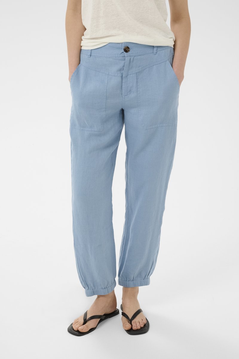 Persona con pantalones azul claro con puños elásticos, camiseta blanca y chanclas negras, de pie con las manos en los bolsillos frente a un fondo liso.