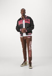 Svart satin bomberjacka med röd "BULLS" text, i kombination med bruna sweatshirtbyxor med "CHICAGO BULLS" tryck, och tvåfärgade sneakers.