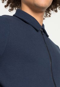 Laivastonsininen vetoketjullinen takki, jossa on teksturoidtu kangas, seisova kaulus ja kapea malli. Kappaleessa on näkyvä vetoketjuaksentti ja sileä viimeistely.