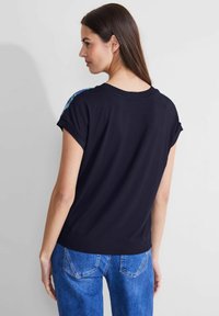Camiseta negra de manga corta con cuello redondo, que presenta acentos florales en los hombros, combinada con jeans de mezclilla azules. Textura suave y lisa.