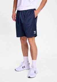 Navy urheilushortsit, joissa on joustava vyötärö, sivutaskut ja valkoiset brändäykset. Yhdistetty valkoisiin sneaker-kengiin ja lyhyisiin sukkiin.