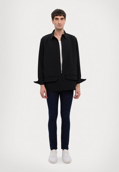 Camicia nera con bottoni, due tasche frontali, maniche lunghe e una vestibilità comoda, abbinata a jeans scuri e sneakers bianche.