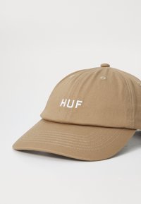 Tan baseball cap lavet af bomuld med en buet skygge, der har et hvidt broderet "HUF" logo på forsiden. Synlige syninger.